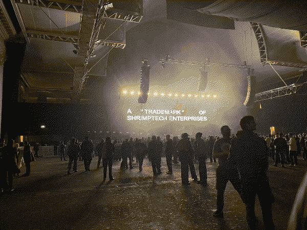 A crowd of people inside a vast dark hall, looking towards a stage. The screen behind the stage says 'A "TRADEMARK" OF SHRIMPTECH ENTERPRISES'