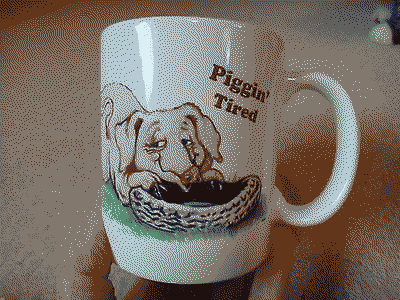 Someone holding up a mug that has a drawing of a smirking pig sprawling over a car tyre, with the text Piggin tired